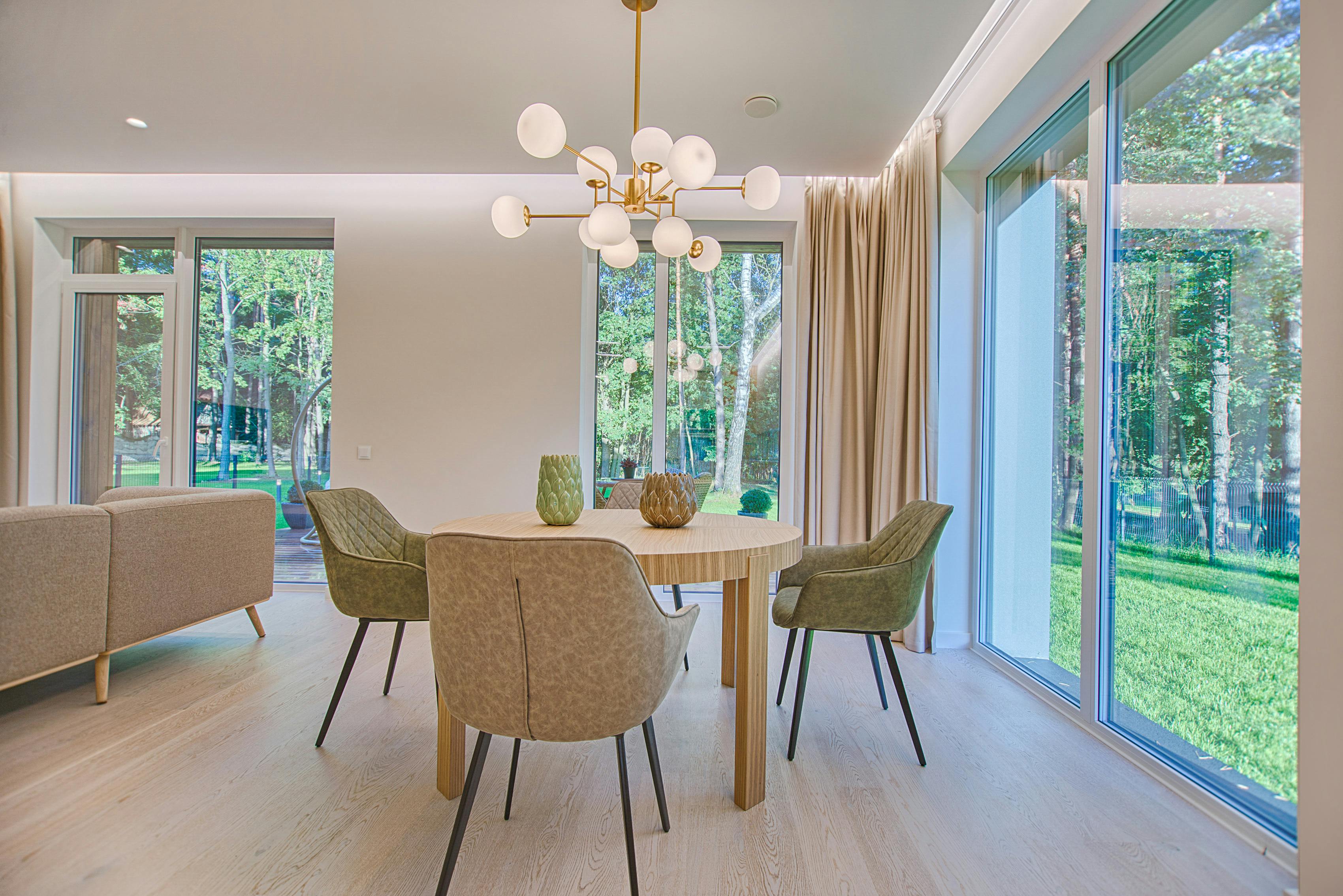 Moderne eetkamer met een ronde houten tafel, vier groene stoelen en een kroonluchter. Grote ramen bieden uitzicht op een weelderige groene tuin.